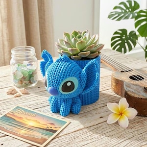 May include: A blue crocheted Stitch planter holds a succulent plant. The planter is next to a postcard with the word "Hawaii" and a ukulele. A jar of sea glass and a flower are also on the wooden surface.