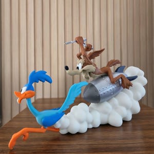 May include: A multi-colored figurine featuring a blue Road Runner bird running in front of a brown Wile E. Coyote riding a silver rocket. The rocket is propelled by a large, white, cloud-like base that extends behind the characters, creating a dynamic scene.