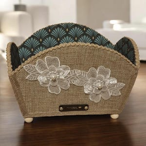 Puede incluir: Una cesta decorativa de arpillera beige con un diseño floral. La cesta presenta dos flores de encaje blanco con centros de perlas. El forro interior tiene un patrón geométrico turquesa y negro. La cesta tiene una pequeña etiqueta marrón que dice "Hand Made".