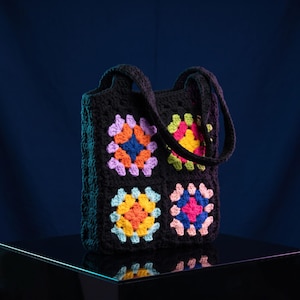 May include: A black crochet tote bag featuring four square panels with colorful granny square patterns in shades of pink, yellow, orange, blue, and light purple. The bag has two black crochet handles and a textured, handmade yarn construction.