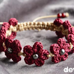 Könnte beinhalten: Handgefertigtes Makramee-Armband mit bordeauxroten Häkelblumen mit silbernen Perlen in der Mitte. Das Armband hat ein beiges Makramee-Band und verstellbare beige Kordeln mit roten Perlen. Auf einer grauen Oberfläche.