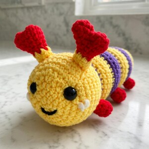 May include: A handmade crochet caterpillar toy in yellow, purple, and red. The caterpillar has a smiling face with black eyes and red heart-shaped antennae. The body is striped with yellow and purple. The toy is made of soft yarn.