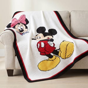 May include: A white crocheted blanket featuring a large graphic of Mickey Mouse in red shorts and yellow shoes, alongside a smaller graphic of Minnie Mouse wearing a pink bow. The blanket has a thick black border with a thin red accent line.