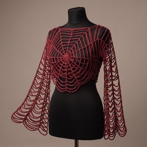 May include: A dark red long-sleeved crop top featuring an intricate, open-knit crochet pattern that resembles a spider web. The garment has a wide, scalloped hemline and flared sleeves, constructed from yarn with a visible, textured stitch design.