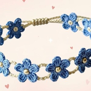 May include: A bracelet featuring a series of blue crocheted flowers connected by a cream-colored cord. Each flower has a small metallic bead at its center. The bracelet includes an adjustable sliding knot closure and blue beads at the ends of the cord.