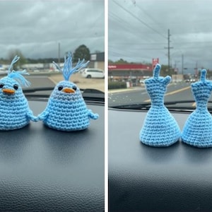 May include: Two blue crochet bird figurines with orange beaks and small wings, and two blue crochet dress figurines. The handmade items are displayed on a car dashboard.