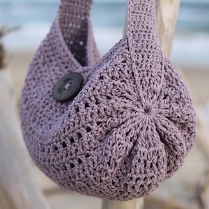 May include: A dusty lavender crochet hobo bag featuring a textured stitch pattern, a single integrated shoulder strap, and a large, dark brown circular button closure centered on the front panel of the rounded, slouchy fabric construction.