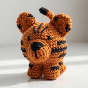 May include: A crocheted tiger toy, featuring orange and black yarn. The tiger has black stripes, small black eyes, and a black nose. The ears and tail are also black. The toy is displayed on a white surface.