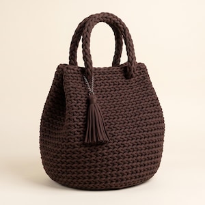 May include: A dark brown crochet handbag featuring a rounded base and a thick, braided handle. The bag is constructed from a chunky yarn with a visible knit texture and includes a matching dark brown tassel charm attached to the handle base.