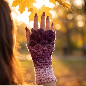 May include: A fingerless glove featuring a textured, scale-like crochet pattern that transitions from a deep dark purple at the top to a lighter mauve pink at the wrist. The item includes a PDF pattern and video tutorial included.