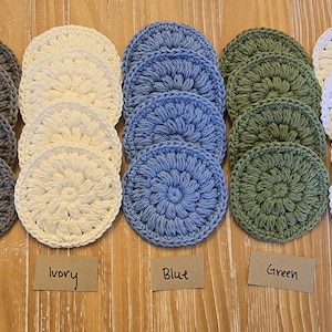 May include: A collection of crocheted coasters in various colors, including salmon, gray, ivory, blue, green, white, and brown. Each coaster is round with a textured, floral-like pattern. The colors are labeled on small brown tags.