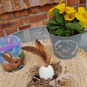 Könnte beinhalten: Ein Ostermotiv mit einem weißen Ei mit Federn in einem Nest, einem Topf mit gelben Primeln und einem Kerzenhalter mit Hasenmotiv. Die Gegenstände sind auf einer gewebten Unterlage angeordnet, mit einer Backsteinmauer im Hintergrund.