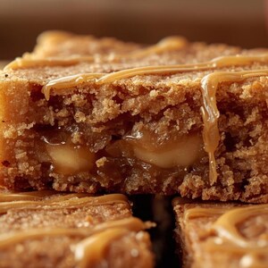 May include: Close-up of a caramel-drizzled dessert bar. The bar has a light brown, textured exterior with a creamy, caramel-filled centre. Caramel sauce is drizzled over the top, with some dripping down the sides. The dessert appears to be freshly baked.