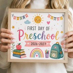 以下が含まれることがあります： First Day of Preschool 2026-2027という文字が書かれた額縁入りのサイン。笑顔の太陽、虹、リンゴが乗った本の山、バックパック、カラフルな旗が手描き風のイラストで描かれています。