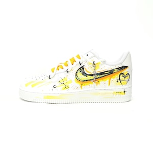 May include: White Nike Air Force 1 sneakers with custom yellow and black paint details. The design includes a yellow swoosh, paint drips, and a heart with a star. The laces are yellow, and a charm hangs from the laces.