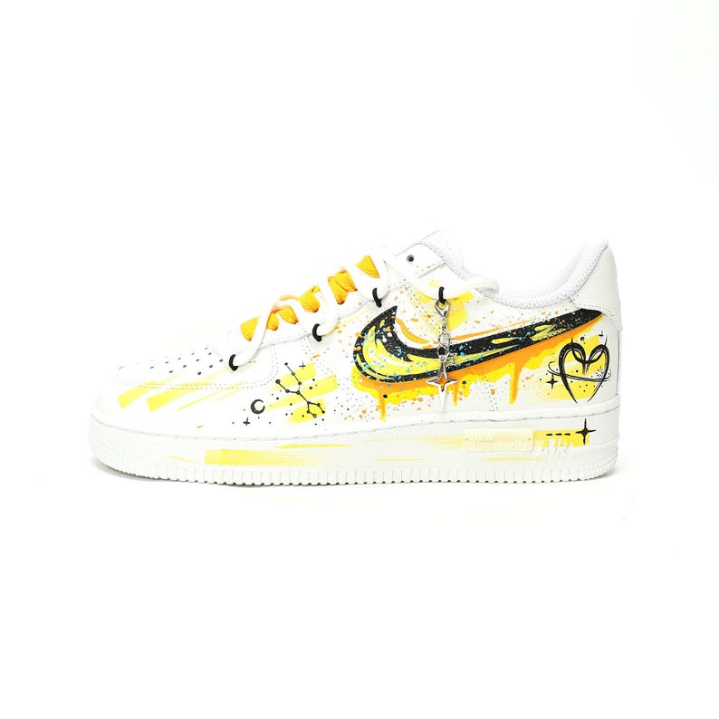 May include: White Nike Air Force 1 sneakers with custom yellow and black paint details. The design includes a yellow swoosh, paint drips, and a heart with a star. The laces are yellow, and a charm hangs from the laces.
