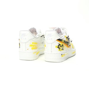 May include: White leather sneakers with yellow and black painted accents. The shoes feature a heart design and the word "AIR" on the heel. The soles are white.