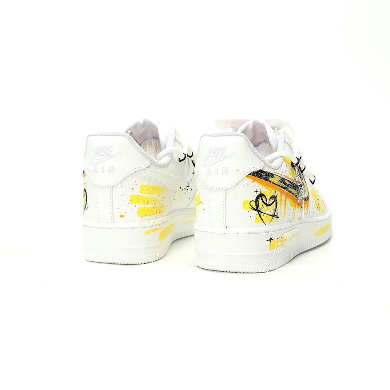 May include: White leather sneakers with yellow and black painted accents. The shoes feature a heart design and the word "AIR" on the heel. The soles are white.