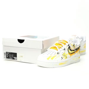 May include: White Nike Air Force 1 Low sneakers with yellow accents and a matching box. The sneakers feature yellow laces, a yellow swoosh, and yellow paint splatter details. The box is white with black text.