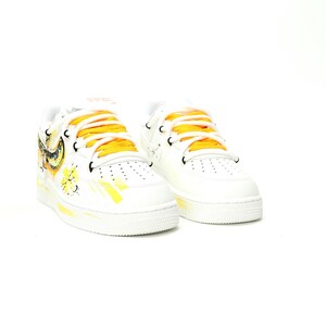 May include: White sneakers with yellow and black accents. The shoes feature yellow laces and a painted design on the side. The design includes a yellow swoosh and other artistic elements. The sneakers have a classic, low-top design.