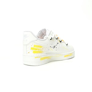 May include: White athletic shoe with yellow paint splatter design. The shoe features a white sole and laces with black accents. The Nike Air logo is visible on the side. A unique, artistic sneaker.