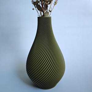 Könnte beinhalten: Eine olivgrüne Vase in Tropfenform mit einer texturierten, spiralförmigen Rippenstruktur an der Außenseite. Die Vase hat einen schmalen Hals, der sich zur Basis hin verbreitert, und enthält ein kleines Arrangement aus getrockneten braunen Pflanzenstängeln.