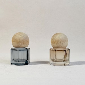 May include: Two small, hexagonal glass bottles with wooden ball-shaped stoppers. One bottle is a smoky gray color, and the other is a light tan. The bottles are displayed against a plain white background.