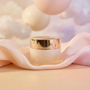 May include: A small, frosted glass jar with a gold-colored lid sits on a draped, light pink fabric. The background features soft, pastel-colored orbs and a cloudy sky, creating a serene and elegant aesthetic. The jar appears to contain a cosmetic product.