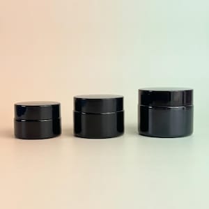 Könnte beinhalten: Drei zylindrische, schwarze Kosmetikdosen in verschiedenen Größen, die in einer Reihe angeordnet sind. Jede Dose hat eine glatte, undurchsichtige schwarze Oberfläche mit einem passenden Schraubdeckel. Die Behälter stehen nebeneinander.