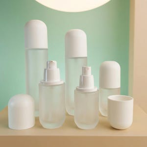 May include: A selection of frosted glass bottles and containers with white lids and dispensers. The bottles are of various sizes and shapes, possibly for skincare or cosmetics. The arrangement is on a neutral-toned surface.