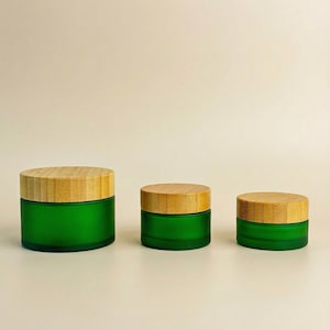 May include: Three green glass jars with bamboo lids are arranged in a row. The jars are cylindrical, with a frosted green finish. The lids are made of light-colored bamboo. The jars are of varying sizes, suggesting a set of containers.