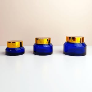 Op de afbeelding: Drie kobaltblauwe glazen potten met goudkleurige deksels zijn gerangschikt op een wit oppervlak. De potten variëren in grootte, met de kleinste links en de grootste rechts. De potten zijn leeg.