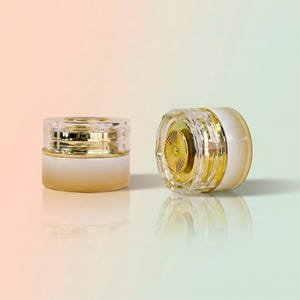 Op de afbeelding: Twee cilindrische cosmetische potjes met een wit en goud kleurverloop aan de basis. Eén potje staat rechtop, terwijl de andere op zijn zijkant ligt, waardoor een goudkleurig, gefacetteerd, rond deksel met een zonnestraalpatroon zichtbaar is.