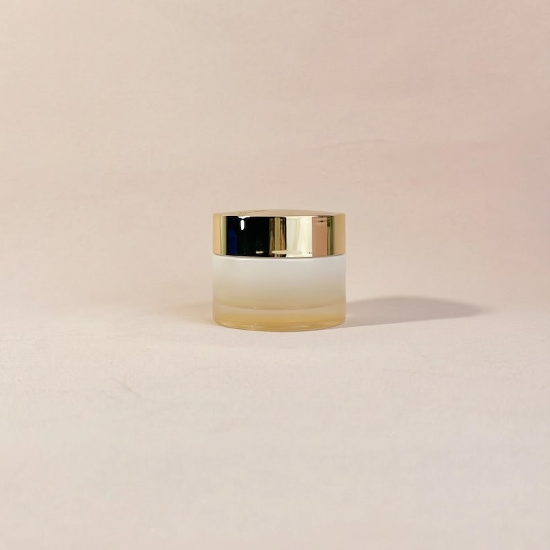 May include: A small, frosted glass jar with a gold-colored lid. The jar has a gradient appearance, transitioning from a light yellow at the base to a white color towards the top. The lid is shiny and reflects light.
