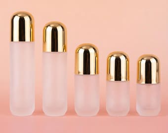 May include: Five frosted glass bottles with gold-coloured caps are arranged in a row. The bottles vary in size, with the tallest on the left and decreasing in height towards the right. The background is a solid light pink.