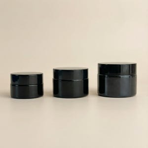 May include: Three black glass jars with matching black lids are displayed. The jars are cylindrical, with a smooth, glossy finish. The sizes vary, with the smallest on the left and the largest on the right.