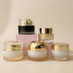 May include: An assortment of cosmetic jars with gold-colored lids and clear or frosted glass containers. The jars are arranged on a light pink surface, showcasing their varying sizes and designs.