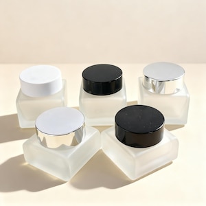 May include: Five frosted glass jars with square bases and rounded tops. The jars have black, silver, and white lids. The jars are arranged on a light-colored surface.