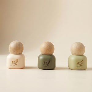 May include: Three decorative bottles with wooden tops. The bottles are in beige, olive green, and light green. Each bottle has a floral design and the word "Luck" printed on the front. The bottles are arranged in a row.