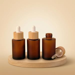 May include: Three amber glass dropper bottles with bamboo lids and white droppers are displayed on a wooden platform. One bottle is open, revealing the dropper. The bottles are cylindrical and have a frosted finish.