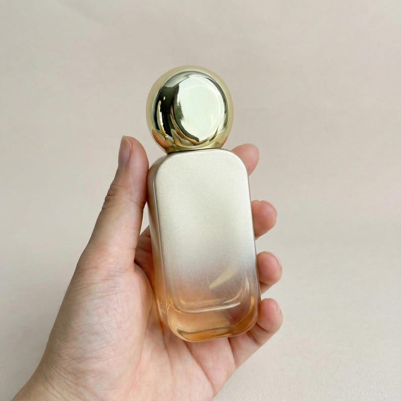 May include: A perfume bottle with a gold-colored spherical cap and a rectangular body. The bottle has a gradient effect, transitioning from a light gold to a peach color. The bottle is held in a hand.
