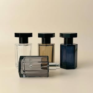 Pode incluir: Quatro frascos de perfume com tampas pretas. Um é transparente, um âmbar e um azul marinho. Um quarto frasco é cinza e está deitado de lado. Os frascos têm formato retangular com tampa preta.
