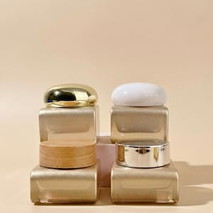Puede incluir: Cuatro frascos cosméticos con diferentes estilos de tapa. Dos frascos tienen tapas doradas, uno con tapa de madera y otro con tapa blanca. Los frascos son cuadrados y tienen un acabado dorado. El fondo es de color beige claro liso.