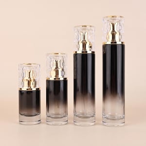 May include: Four black and clear glass cosmetic bottles of varying sizes. Each bottle has a gold-colored pump and a clear, faceted cap. The bottles have a gradient effect, transitioning from black to clear.