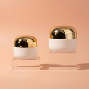 Könnte beinhalten: Zwei weiße und goldene Kosmetikgläser in verschiedenen Größen werden auf transparenten Acrylblöcken vor einem pfirsichfarbenen Hintergrund präsentiert. Die Gläser haben eine abgerundete Form mit weißem Boden und glänzendem Golddeckel.