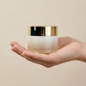 May include: A frosted glass jar with a gold-colored lid. The jar is held in an open hand against a neutral background. The jar appears to be empty and is likely used for cosmetics or skincare products.
