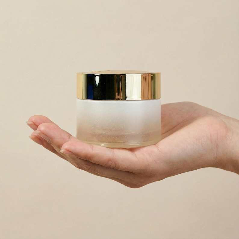 May include: A frosted glass jar with a gold-colored lid. The jar is held in an open hand against a neutral background. The jar appears to be empty and is likely used for cosmetics or skincare products.