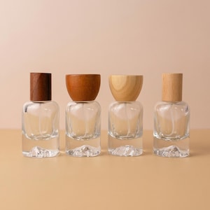 May include: Four clear glass perfume bottles with wooden caps. The caps vary in color from dark brown to light tan. The bottles are arranged in a row against a neutral background.