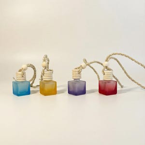 Pode incluir: Quatro pequenas garrafas quadradas de vidro em azul, amarelo, roxo e vermelho, cada uma com uma tampa de madeira e um cordão tecido para pendurar. As garrafas foscas são provavelmente ambientadores ou artigos decorativos.