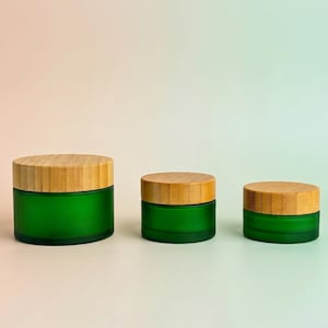 Può includere: Tre barattoli cilindrici di dimensioni diverse, ciascuno caratterizzato da una base in vetro smerigliato verde smeraldo e un coperchio in bambù testurizzato di colore chiaro. I barattoli sono disposti in fila dal più grande al più piccolo.